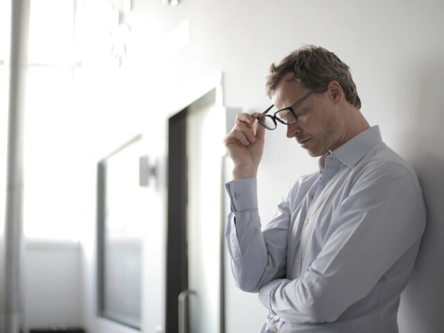 Man leaning on wall holding glasses looks tired