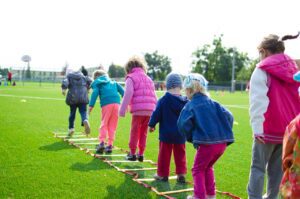 Kids playing game lining up on green grass jumping over ladder hurdles