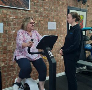 Lady exercising on bike in gym with exercise physiologist talking to her